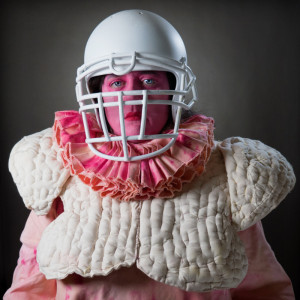 A person in a theatrical costume with a stark contrast of elements: their face is painted pink, and they are wearing a white football helmet with a face cage. The costume includes a padded, quilt-like garment with ruffled accents, combining elements of traditional Elizabethan attire with modern protective sports gear. 