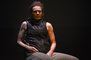 A person wearing a black puffer vest and khaki pants, sitting and looking down, with sweat and a bandage visible on their arm, under a spotlight in a dark setting.