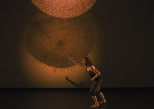 A barefoot performer in a black dress, crouched and reaching forward, with large, circular woven patterns illuminated on the wall behind them.