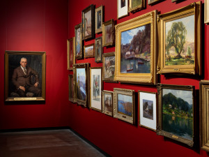 A wall in an art gallery with a variety of framed artworks against a red background.