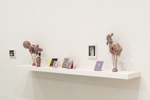 A shelf display against a white wall featuring whimsical sculptures covered in colorful patterns, and assorted books.