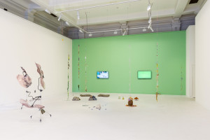 An art installation featuring abstract sculptures on floor stands, strings of colorful decorations hanging from the ceiling, and two video works on a green wall inside a gallery with bright overhead lighting.