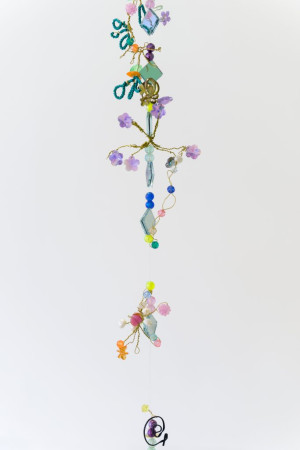 A vertical, hanging mobile composed of colorful beads, faux flowers, and various whimsical elements strung together, creating an eclectic decorative piece against a white background.