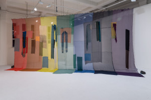 A large-scale, multi-colored fabric installation with various hanging textiles in different shapes and shades, creating a layered, curtain-like effect in an art gallery space.