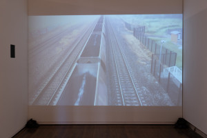A video projection on a wall showing an aerial view of a long freight train traveling on parallel railway tracks in a rural area.