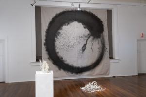 An art installation in a gallery featuring a large canvas with a black circular design on the wall, a pedestal with a small white sculptural piece, and a pile of white material on the wooden floor.