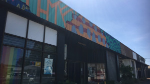 A modern building featuring large windows, with a colorful, abstract mural on its upper façade under a clear blue sky.