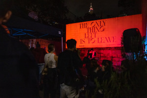 A night scene with a DJ under a sign that reads, THE ONLY THING LEFT IS TO LEAVE, with an audience and Sydney-city lights in the background.