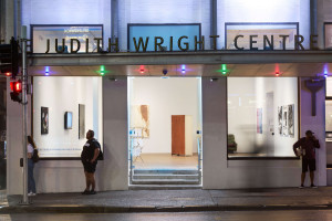 The illuminated façade of the Judith Wright Centre at night, with its name in bold letters above the entrance. Artworks are visible through the glass front, and three individuals stand outside, each engaged in different activities: one walking, another observing, and the third on a phone.