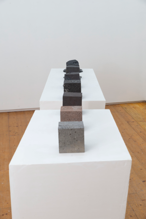 A series of seven textured blocks in shades from dark to light gray are aligned on two connected white pedestals on a wooden floor, creating a gradient effect.