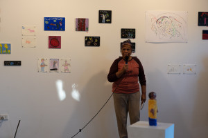 A person is speaking into a microphone in an art gallery, with a collection of various drawings displayed on the wall behind them and a small sculpture displayed on a plinth.