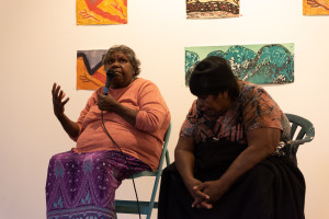 Two people seated on chairs at an event; one is actively speaking into a microphone with an expressive gesture, and the other is seated beside, looking downward. Artwork is displayed on the wall behind them.