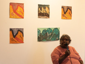 A person sits speaking into a microphone against a gallery wall adorned with five colorful paintings.