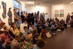 A diverse audience seated and standing in a packed art gallery, clapping and focused on something outside of the frame. The gallery walls are adorned with various artworks.