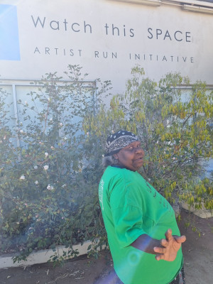 A person stands in front of a building with the text 'Watch this SPACE: ARTIST RUN INITIATIVE' on the wall. The individual is wearing a bright green shirt and a bandana, extending their hand toward the camera, against a backdrop of leafy greenery.