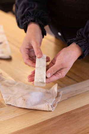 Close-up of hands holding a piece of white, semi-translucent marble or stone, with a cloth pouch partially visible on a wooden surface. The focus is on the texture and quality of the material being examined.