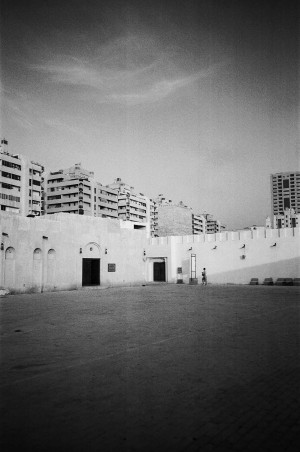 A black-and-white photograph captures an open courtyard in Sharjah, UAE, known as Calligraphy Square. The foreground features a spacious, paved area with a single person walking near the entrance. The surrounding walls are tall and white, with traditional arched doorways and minimalistic detailing. In the background, modern residential buildings rise above the courtyard, contrasting with the traditional architecture of the square. The sky above is mostly clear with some wispy clouds, adding depth to the scene. The image highlights the blend of old and new in the urban landscape.