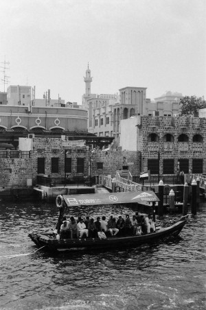 A black-and-white photograph shows a traditional wooden abra boat filled with passengers crossing a waterway in Dubai. The boat has a canopy with the words Dubai and RTA printed on it. In the background, there is a mix of older buildings made of stone and modern structures, including a minaret and various architectural details that reflect the region's heritage. The scene captures a busy and culturally rich area, highlighting both the historical and contemporary elements of the city.