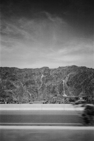 A black-and-white photograph shows a view of a mountainous landscape taken from a moving vehicle. In the foreground, a highway guardrail and the blurred image of a speeding motorcycle are visible. The rugged, rocky mountains dominate the background, with sparse vegetation and a tall, thin structure, possibly a flagpole, standing out against the natural terrain. The sky above is partly cloudy, adding depth to the scene. The image captures the contrast between the motion of the vehicle and the stillness of the mountainous environment.