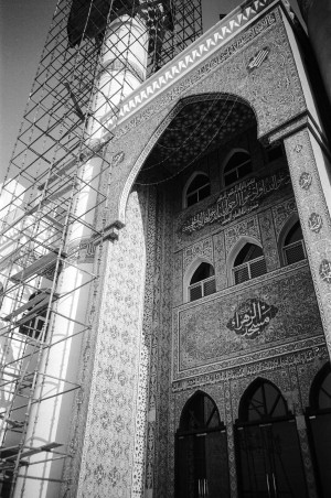 A black and white image, captured on 35mm film by Gillian Karyooz in 2023, features the front entrance of the Al Zahra Mosque in Sharjah, UAE. The façade is adorned with elaborate patterns and Arabic calligraphy. Scaffolding is set up on the left side, signifying ongoing maintenance work. The mosque is located in Al Muraijah Square, part of the Al Mureijah historical quarter known for its historical significance dating back to the nineteenth century.