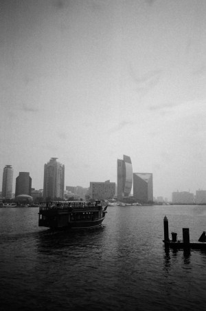 A black-and-white photograph depicts a view of Dubai Creek with modern skyscrapers in the background. In the foreground, a traditional wooden boat is sailing on the water, creating a stark contrast between the old and new. The skyline features several distinctively shaped buildings, including one with a unique, angled design. The waterway is calm, and the image captures the blending of cultural heritage with contemporary urban development in Dubai.