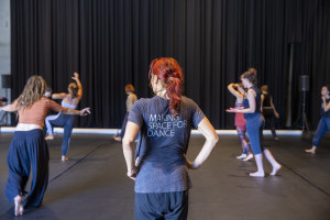 A dance class in session with several participants moving in various poses. A person with red hair and a ponytail, is in the foreground, wearing a gray shirt that says “MAKING SPACE FOR DANCE.” The background features a black curtain and minimalistic setup.