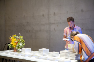 Two people are examining books on a table covered with stacks of white books. One person is flipping through a book, while the other is bending over to read. A vase of colorful flowers is also on the table, and the background is a plain concrete wall.