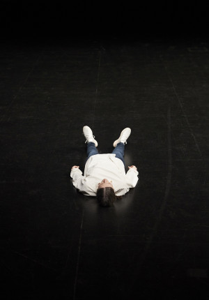 A person in a white hoodie and blue jeans lies flat on their back on a dark stage, arms relaxed by their sides, with their feet pointed towards the camera.