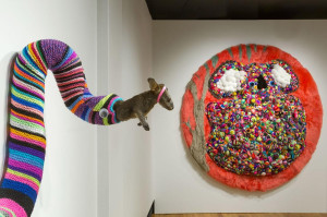 An art installation with a stuffed animal wearing a pink band leaping from a long, multicolored knit tube attached to the wall. To the right, a large, circular furry sculpture with red, grey, and white sections is filled with colorful, beaded textures, in the shape of a koala.