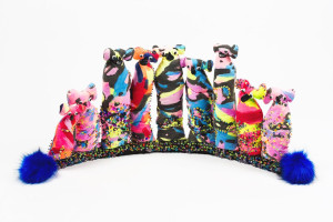 row of colorful, stylized animal figures with pompom details and vibrant patterns against a white background. The animals are adorned with sequins and beads, and there are large blue pompoms at both ends of the row.