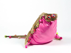 A whimsical sculpture of an animal with a shiny pink body, a fur-trimmed back, and twig-like protrusions adorned with colorful pompoms, against a white background.