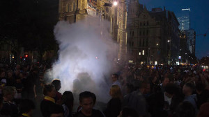 A crowd of people on a city street at dusk, enveloped in smoke from a source obscured from view.