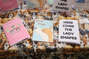  An assortment of printed materials, including zines, laid out on a fabric surface with a repetitive pattern of various dog faces. Central are three books with prominent titles: The Gang's All Queer, Good Dogs, and HERE COME THE LADY SHAPES.