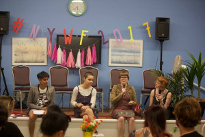  Four panelists are seated in front of an audience at a cultural event, with a hashtag #NYWF2017 displayed in large, colorful letters above them.