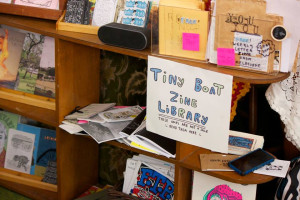 A wooden bookshelf displaying an eclectic collection of zines and small books, labeled as the Tiny Boat Zine Library. A handwritten sign on the shelf states THESE ONES ARE NOT FOR SALE - READ THEM HERE, inviting readers to browse on-site.