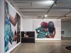 Spacious gallery interior showcasing a large photograph of two people with their backs turned, beside a monitor and another large photograph of the same two people.