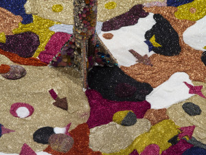 Detail of a glittery, colorful artwork with sequins and beads creating an abstract pattern, with a central figurative element resembling a tree trunk.
