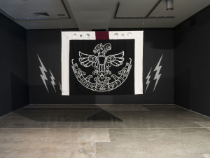Art installation displaying a black textile banner with a white ornate pattern resembling a crest, accented with lightning bolt designs on a black wall background.