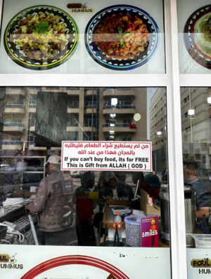 A storefront window displays a sign with text in Arabic and English that reads: If you can't buy food, it's for FREE. This is Gift from ALLAH (GOD). The window also shows images of various dishes, including foul and hummus. Inside the store, a person is visible working behind the counter, and some customers are seated at tables. The reflection on the glass shows buildings across the street.