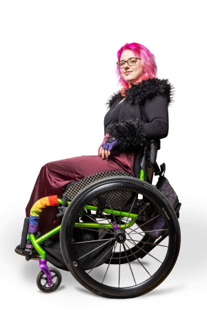 Robin with their wheelchair in full profile against a blank background, smiling slyly at the camera.