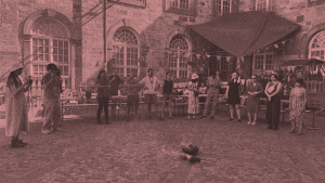 A group of people stand in a circle in a courtyard. It is daytime but a fire is in the centre of them.